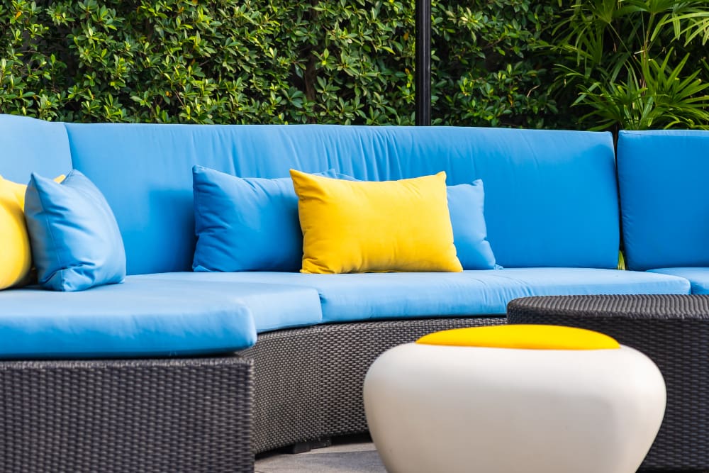 A curved outdoor wicker sofa with blue cushions and yellow accent pillows sits by a round white table with a yellow top, set against a green hedge—ideal for relaxing after your Leather garment cleaning in Syracuse.