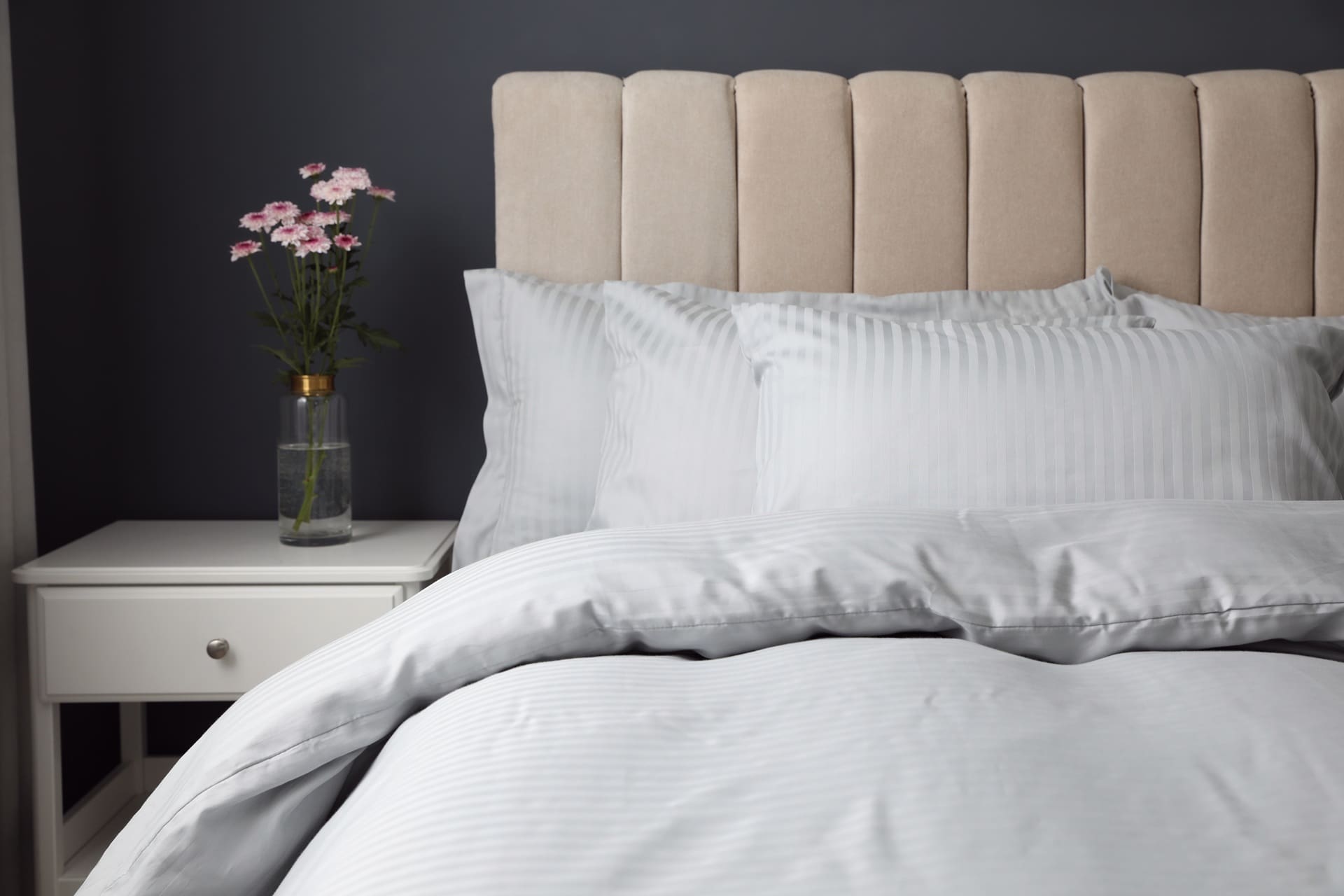 A neatly made bed with light gray bedding and a beige headboard sits beside a white nightstand holding pink flowers, set against a dark gray wall—perfect for relaxing after your wedding dress cleaning in Baldwinsville.