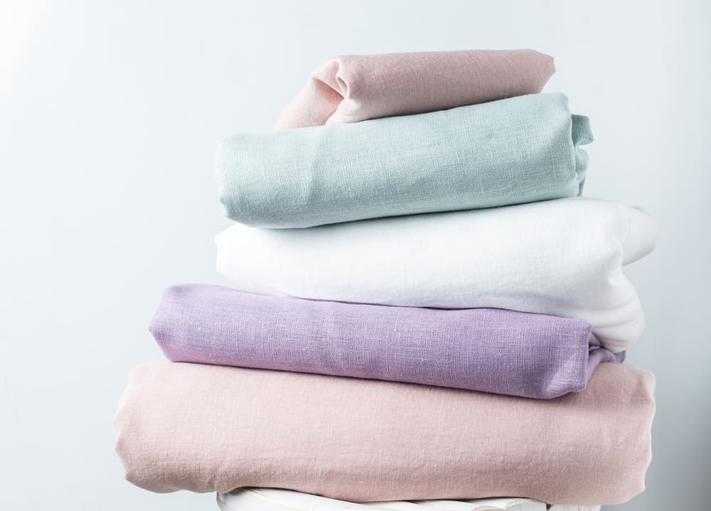A stack of four folded linen fabrics in pastel colors—pink, lavender, white, and light green—on a white surface against a light background, ideal for showcasing the quality care found with wedding dress cleaning Baldwinsville.