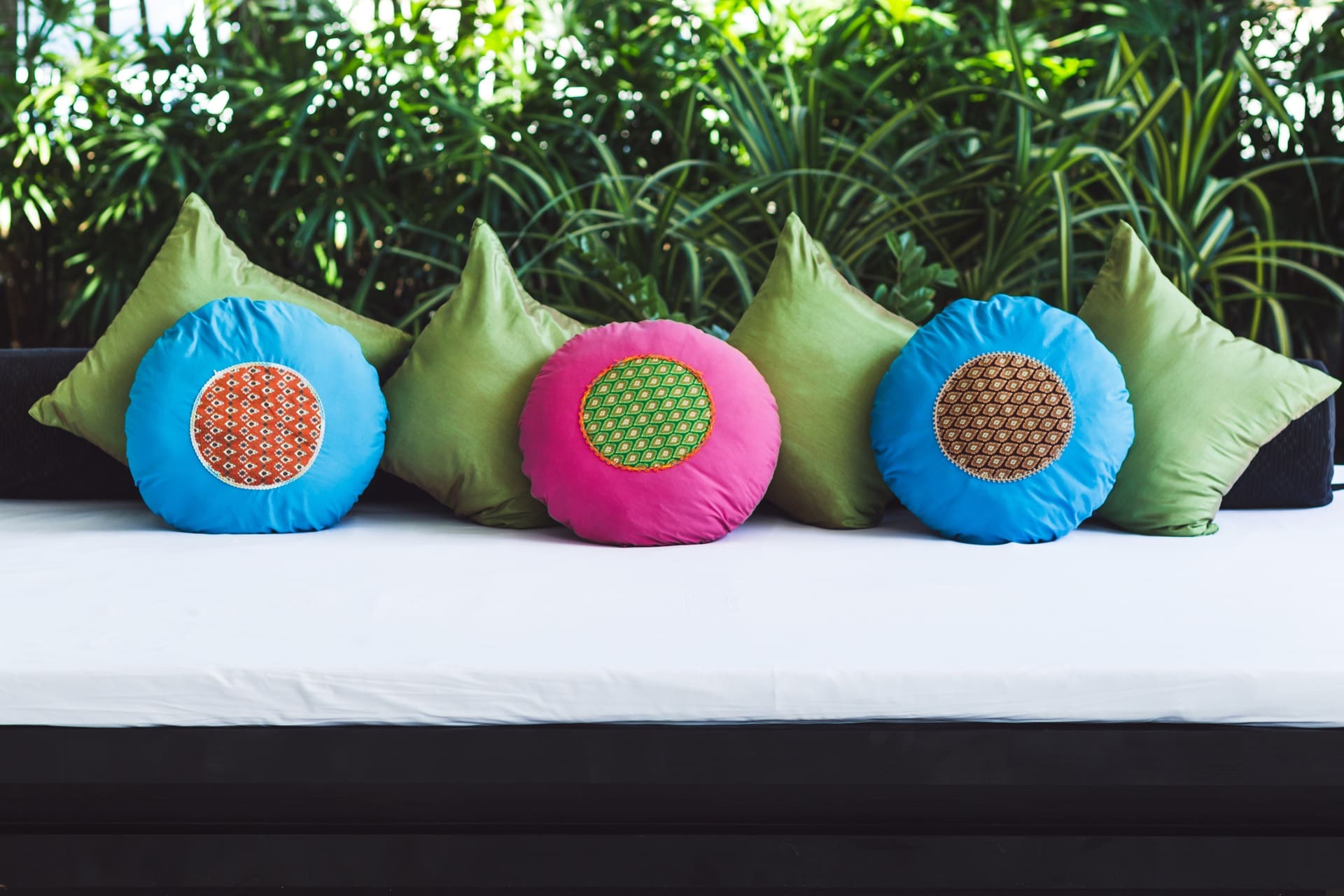 A white cushioned bench with four green triangular pillows and three round pillows—two blue and one pink—arranged against a backdrop of green plants, ideal for relaxing after a day of leather garment cleaning in Manlius.