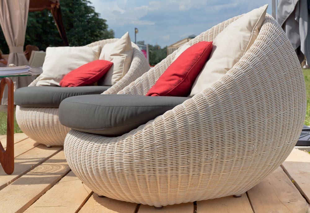 Two round wicker chairs with gray cushions, beige pillows, and red accent pillows are placed on a wooden deck outdoors, the perfect spot to relax after a visit for leather garment cleaning in Syracuse.