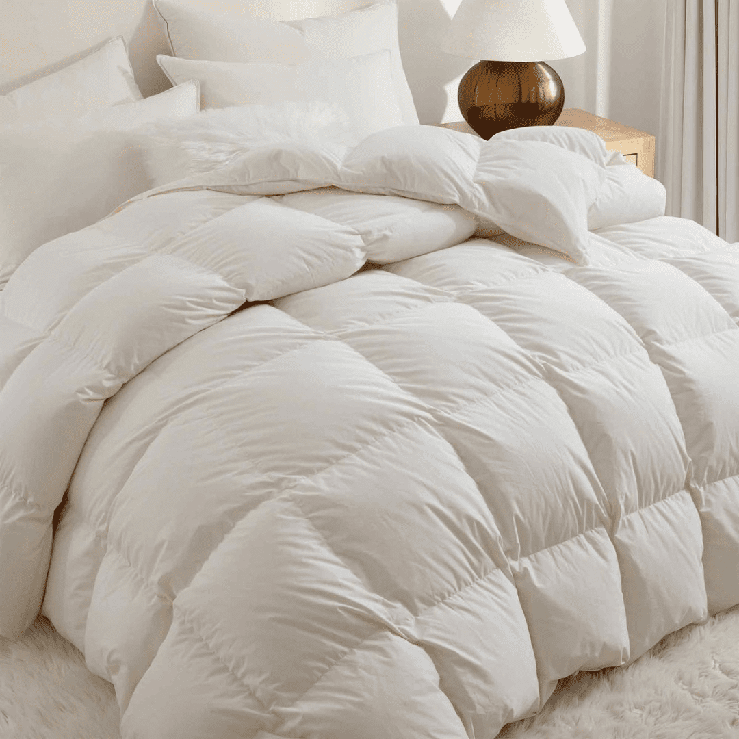 A neatly made bed with a white, puffy comforter and multiple pillows beside a wooden nightstand with a lamp.
