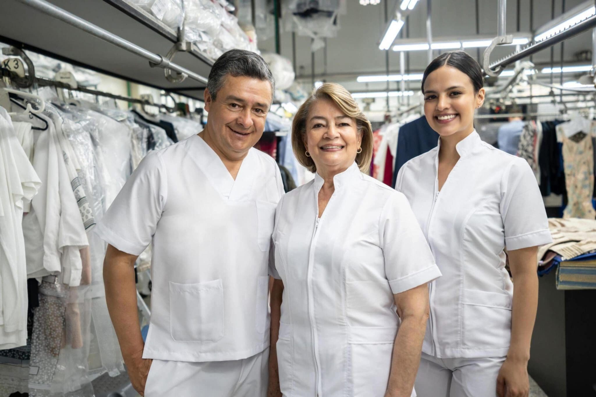 Three people in white uniforms stand and smile in a laundry or dry cleaning facility, surrounded by hanging clothes and industrial equipment.