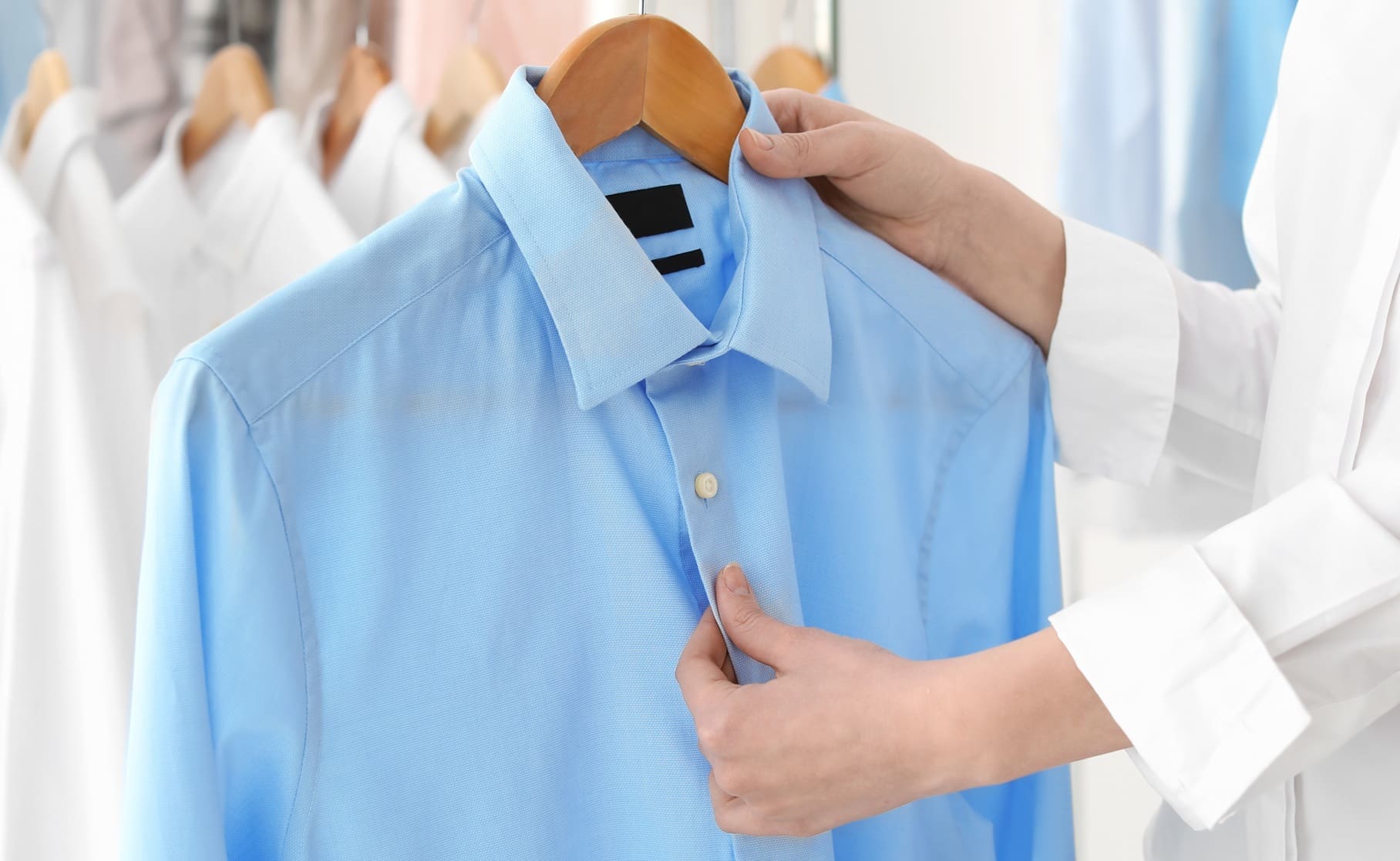 A person holds a light blue dress shirt on a wooden hanger, inspecting or buttoning it, with other shirts hanging in the background.