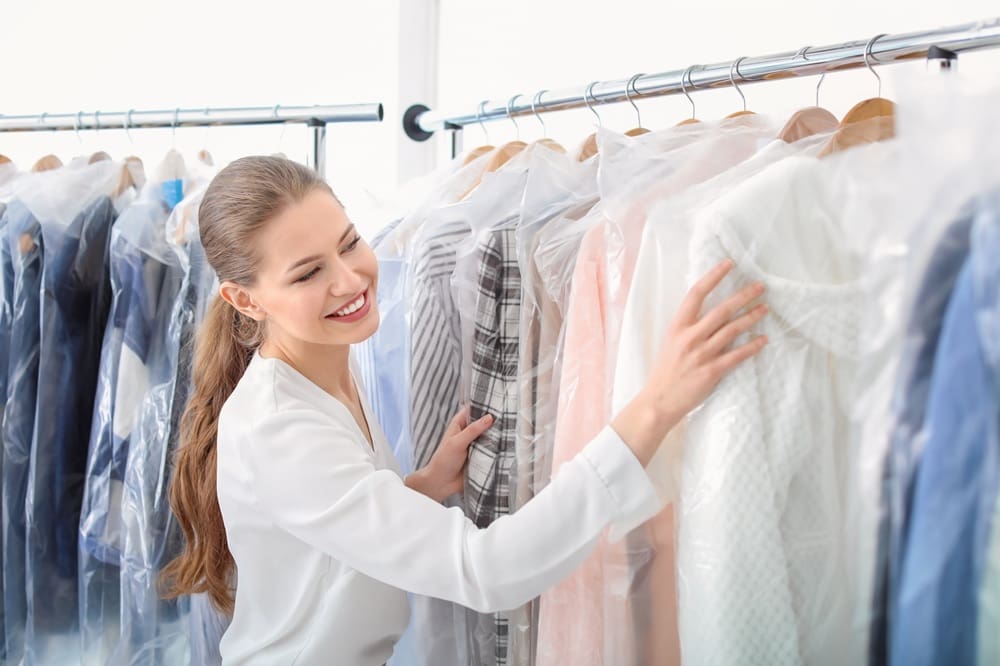 A woman smiles while selecting clothes covered in plastic on hangers from a rack, possibly at a dry cleaner or laundry service.