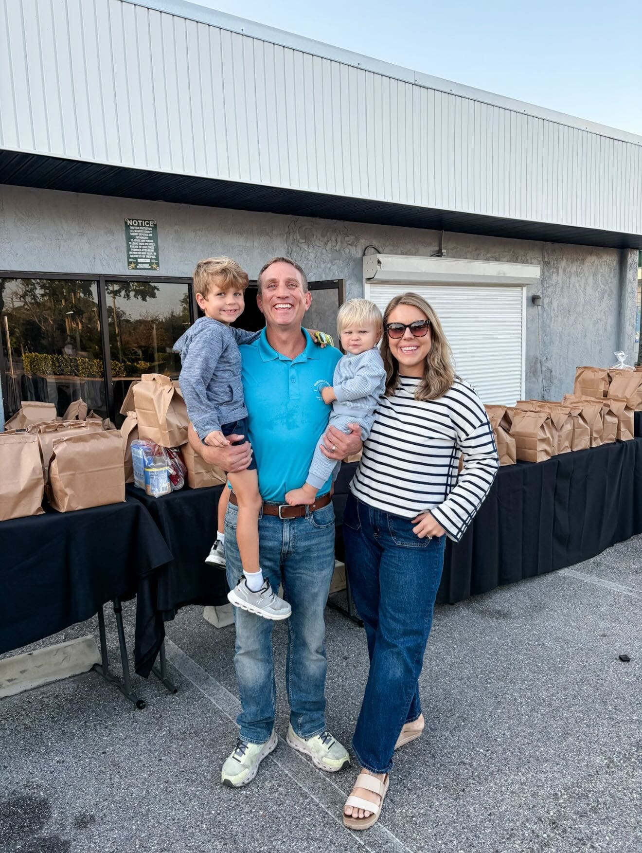 A family of four stands in front of a table with many brown paper bags outside a building; the parents are smiling and holding their two young children.