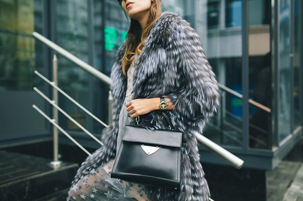 A person wearing a gray faux fur coat and holding a black handbag stands in front of a modern glass building.