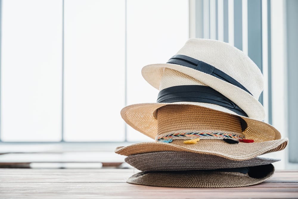 Three straw hats stacked on a wooden surface, with a bright window and blurred vertical blinds in the background.