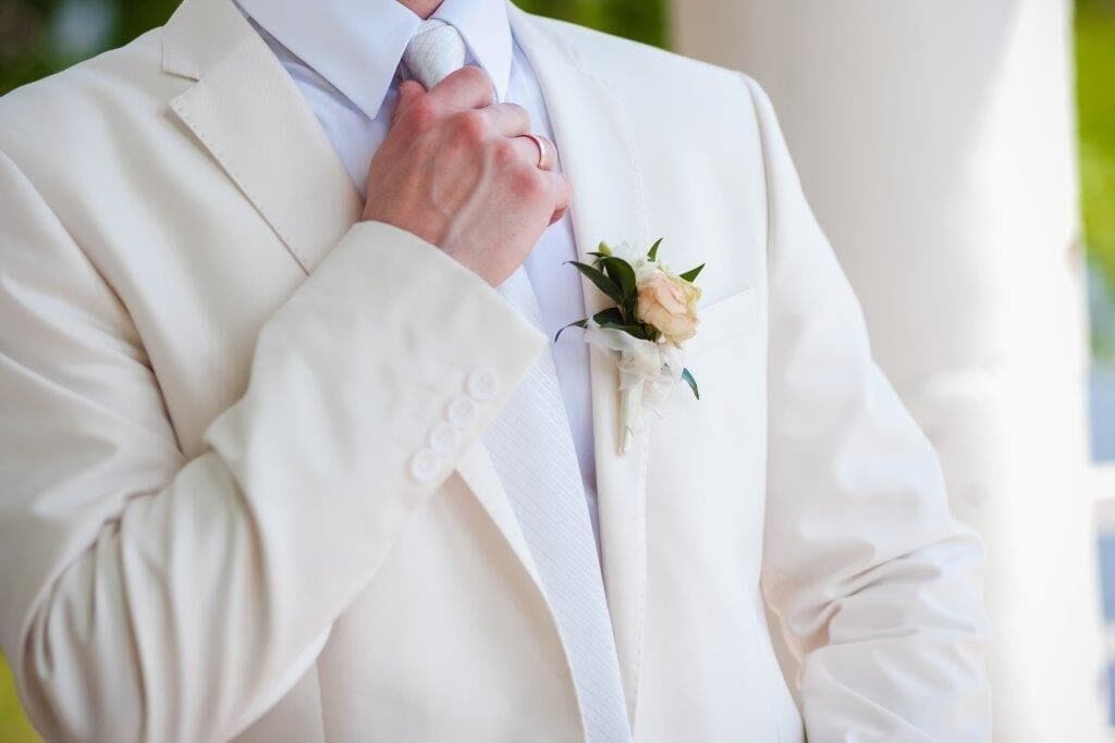 A person in a white suit adjusts their tie, wearing a boutonniere with a light peach flower pinned to the lapel.