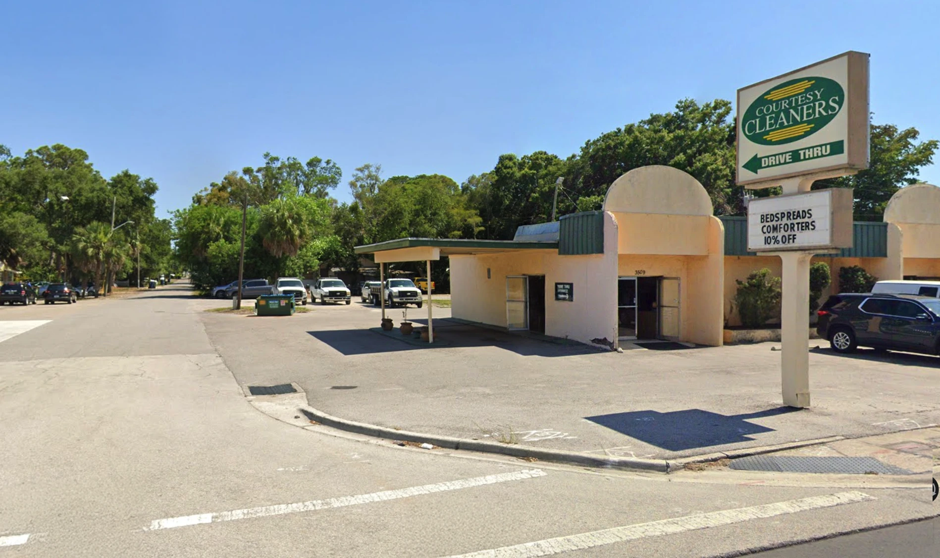 A small dry cleaners building with a drive-thru lane and parking lot stands on a corner, with cars parked nearby and trees in the background.