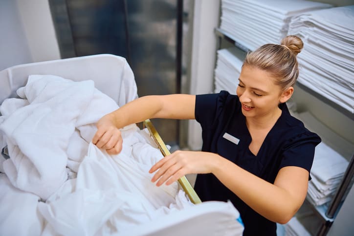 A woman in uniform sorts white linens in a laundry room with shelves of folded towels in the background.