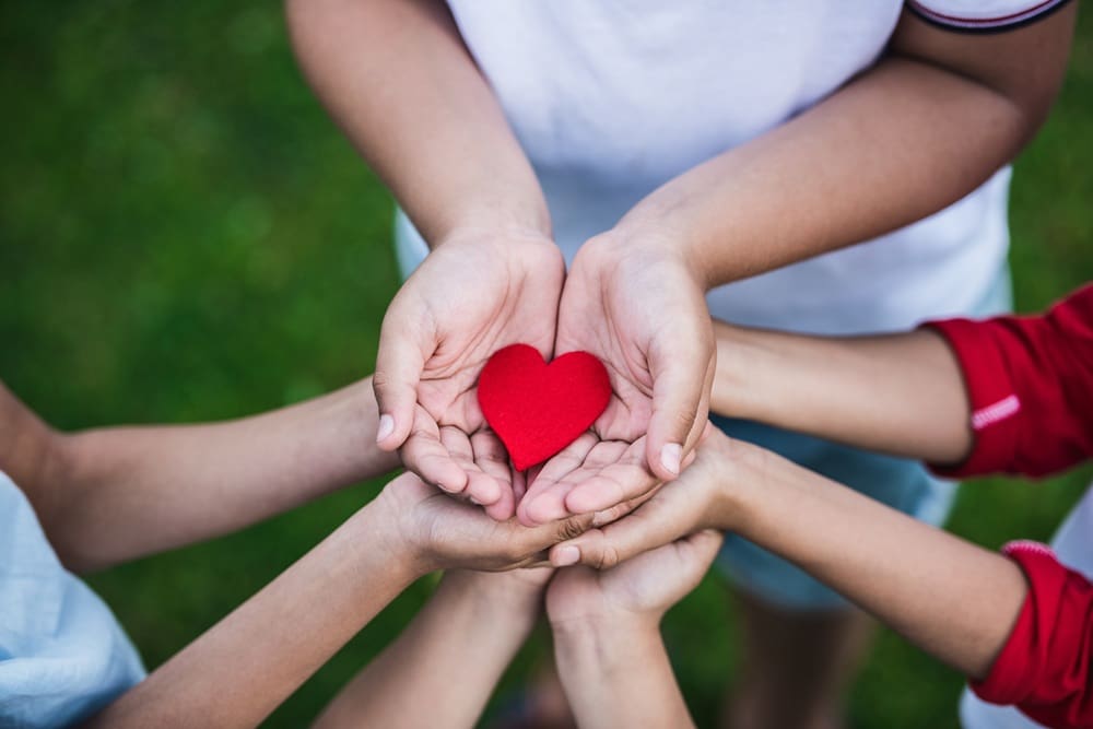 Several children’s hands are cupped together, holding a small red heart shape, with green grass in the background.