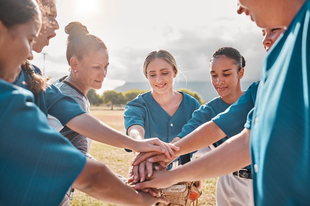 A group of women in blue baseball uniforms stand in a circle stacking their hands together outdoors on a field.