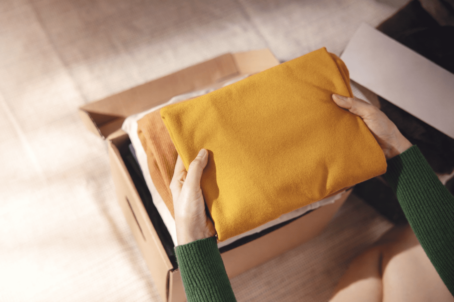 A person holding a folded yellow garment above an open cardboard box containing clothes.