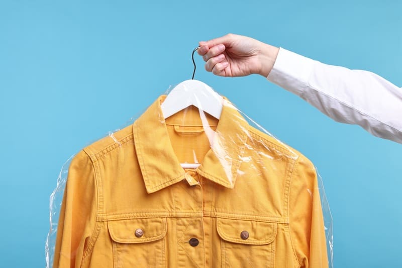 A hand holds a hanger with a yellow denim jacket covered in clear plastic, against a blue background.